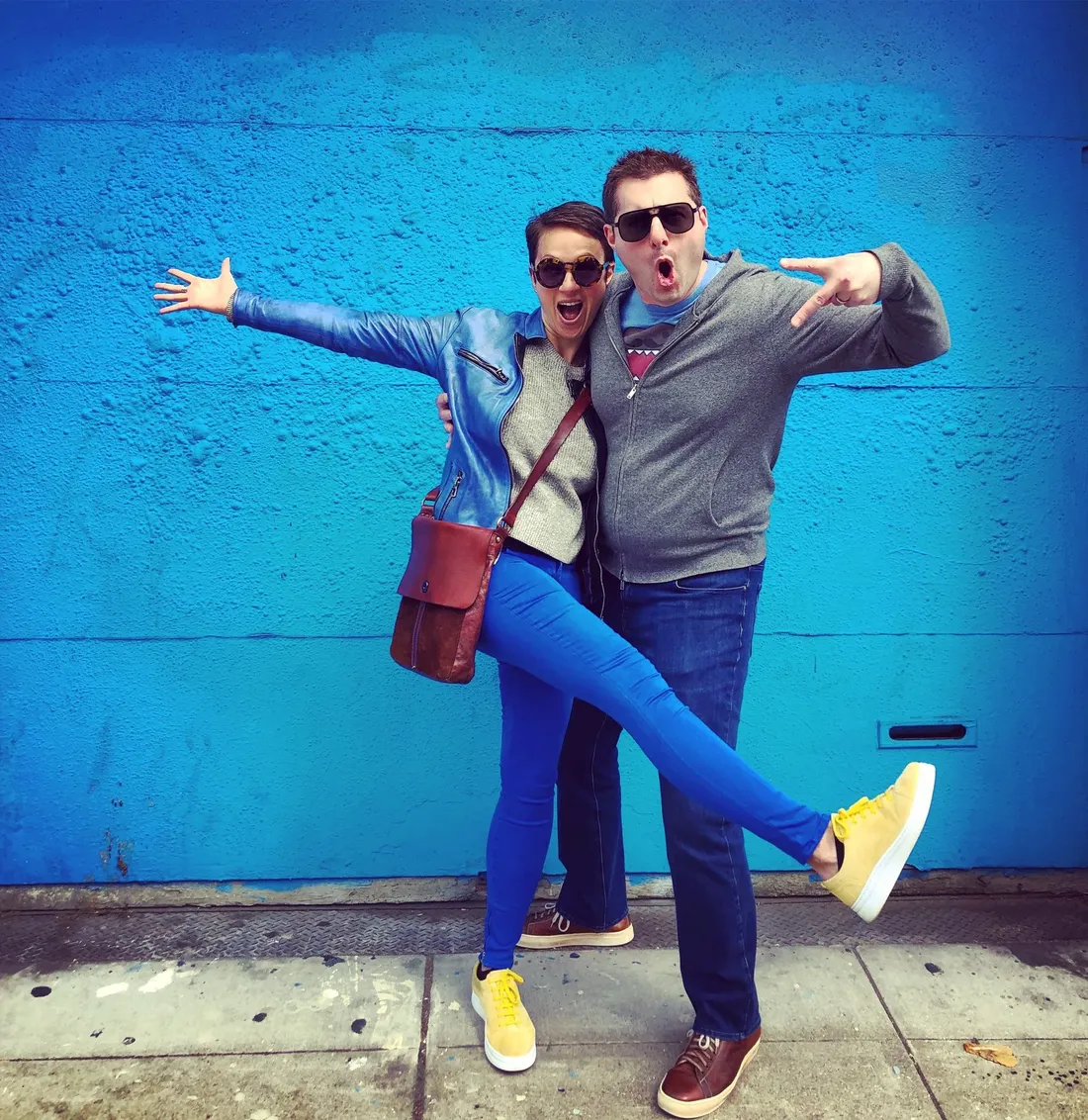 Nataly and Avi in front of a bright blue wall, Nataly with her arms flung wide and one leg kicked out, Avi grinning beside her
