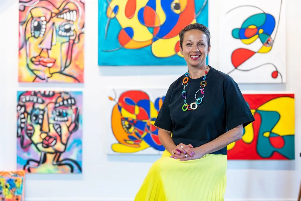 Nataly Kogan smiling, seated in front of a wall of her colorful paintings